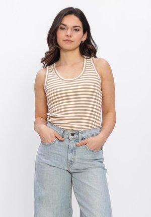 Jonge vrouw met donker haar, die een beige-wit gestreept mouwloos topje en lichtblauwe hooggetailleerde jeans draagt, met de handen in de zakken.