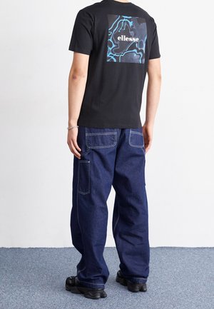 Camiseta de algodón negra con un gráfico azul y gris en la parte de atrás, combinada con pantalones vaqueros oscuros de corte holgado que tienen bolsillos de parche.