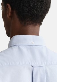 Világoskék ing szabványos gallérral, apró gombdísszel a hátoldalon és pliszírozott yoke-kal a jobb tartás érdekében.