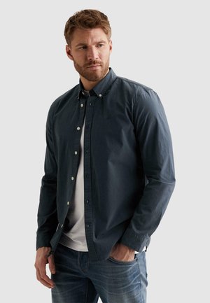 Man met baard, gekleed in een donkerblauw overhemd over een wit T-shirt en blauwe spijkerbroek, staand met één hand in de zak, kijkt zijwaarts.