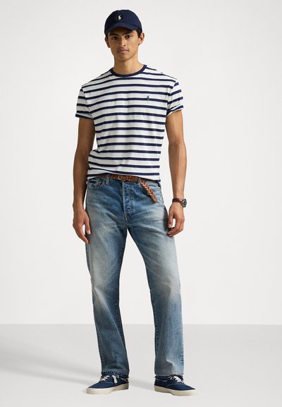 Randig t-shirt i marinblått och vitt, blå jeans, marinblå keps och tygskor. Har en logotyp på t-shirten och ett mönstrat bälte.