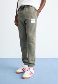 Persoană îmbrăcată în pantaloni de trening Columbia de culoare verde închis, cu talie elastică și adidași albi cu dungi roșii, stând pe un podea de un albastru deschis.
