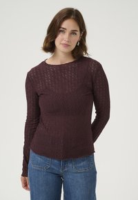 Haut en dentelle à manches longues de couleur bourgogne foncé, avec un col rond et de délicats motifs floraux. Associé à un jean bleu.