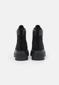 Zwarte enkelbotten van leer en suède, met een chunky zool, een trekband en een geperst logo op de achterkant. Glad en gestructureerd oppervlak.