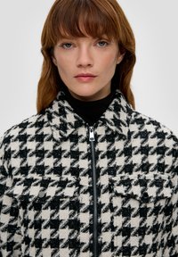 Mustvalge houndstooth mustriga jakk, millel on sulguri esiosa, kaks rinna taskut ja kraega kaelus. Tekstureeritud kangas.