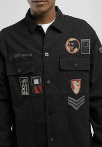 Sort knapjakke med flere broderede mærker, herunder militære insignier og designs i forskellige farver.
