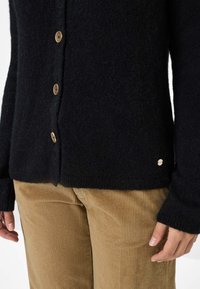 Cardigan nero lavorato a maglia in materiale morbido, con bottoni dorati, vestibilità morbida e polsini a costine. Abbinato a pantaloni in velluto a coste color sabbia.