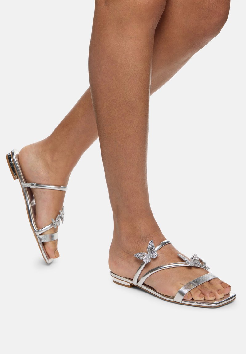 Silver flat sandals with multiple straps, butterfly accents, and a low profile. The upper features a smooth metallic finish and rhinestones.