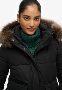 Sort pufferjakke med en faux fur hætte, der har lynlås og sidelommer; båret over en grøn ribbet turtleneck sweater.