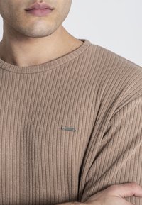 Hnedé rebrované úpletové tričko s dlhým rukávom, okrúhlym výstrihom a malou striebornou nášivkou značky na ľavej hrudi. Mäkká textúra a priliehavý strih.