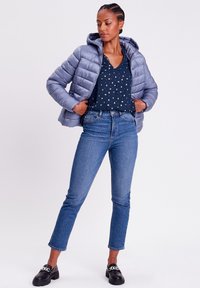 Puffjacke in Hellgrau mit Kapuze, kombiniert mit einer navyblauen Bluse mit weißen Punkten und hochgeschnittenen blauen Jeans. Schwarze Loafers mit Kettendetail.