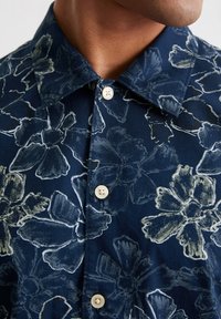 Marineblauwe blouse met bloemenpatroon in lichtbeige. Bevat een klassieke kraag en vier lichtgekleurde knopen aan de voorkant.