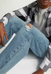 Jean déchiré bleu clair avec des genoux effilochés, en denim. Assorti à une chemise à motifs noir et blanc et à un T-shirt gris en dessous.