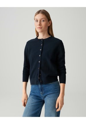 Femme aux longs cheveux châtain clair portant un cardigan noir boutonné et un jean bleu, debout devant un fond clair uni.