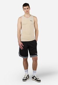 Beige ribbattu toppi, jossa on musta logo, yhdistetty mustiin shortseihin, joissa on valkoiset sivuraidat. Mallilla on päällään mustat ja valkoiset lenkkarit.