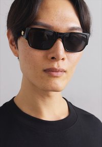 Occhiali da sole neri rettangolari con lenti scure e accessori in oro discreto. Indossati da una persona in una maglietta nera a collo rotondo, vista ravvicinata.