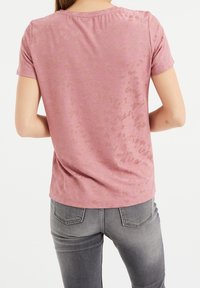 Růžové tričko s krátkým rukávem, které má jemný texturovaný vzor, kulatý výstřih a volný střih, v kombinaci se šedými džínami.