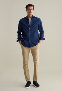Marineblauw overhemd met knoopsluiting, beige broek en donkere sneakers. Het overhemd heeft opgestroopte mouwen; de broek heeft een gladde textuur en een tapered fit.