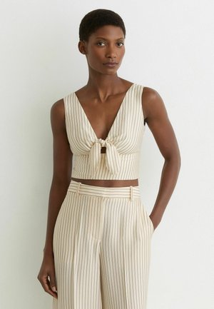 Femme portant un crop top beige à fines rayures avec un nœud à l'avant et un pantalon taille haute assorti, debout, une main dans la poche, contre un mur blanc.