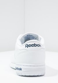 Baskets Reebok en cuir blanc avec un col rembourré, une semelle texturée et un logo bleu marine sur le talon et le côté. Design minimaliste.