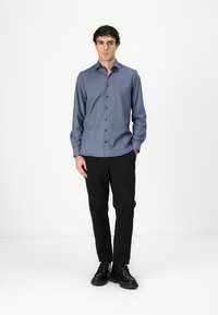 Cămașă albastră cu mâneci lungi, guler cu nasturi și nasturi frontali, asortată cu pantaloni negri și pantofi negri, pe un fundal alb.