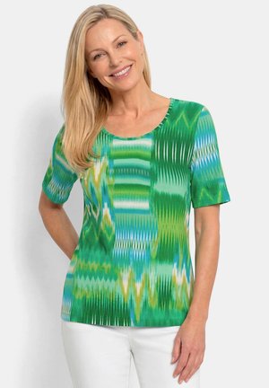 Femme souriante aux longs cheveux blonds portant un haut vert à manches courtes à motifs et un pantalon blanc, debout devant un fond clair uni.