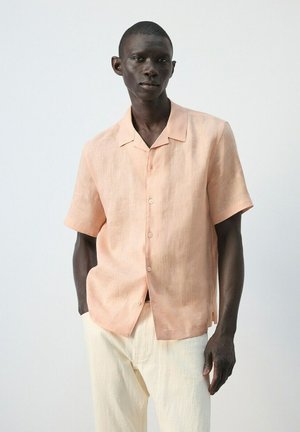Homme à la peau foncée portant une chemise à manches courtes couleur pêche clair boutonnée et un pantalon blanc cassé, debout avec une main dans la poche sur un fond uni.