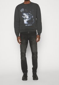 Sudadera negra de cuello redondo con un gráfico de un lobo y estrellas, combinada con jeans negros con acentos de rodillas acanaladas y zapatillas negras.