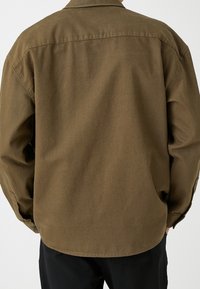 Camicia button-up in cotone marrone con vestibilità rilassata, cuciture visibili e orlo curvo. Il tessuto appare testurizzato e resistente, con un design semplice.