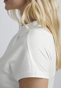 Hvid polo-shirt lavet af struktureret stof, med høj krave og korte ærmer, vist fra siden med glat, lige blond hår.