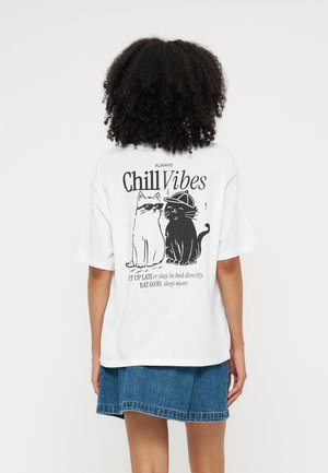 Personne avec des cheveux bouclés porte un t-shirt blanc avec des illustrations de chats en noir et blanc et un texte sur une ambiance détendue, associé à une jupe en jean bleue.