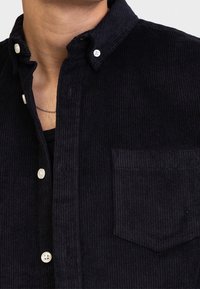 Camisa de pana color azul marino con collar tipo button-down, bolsillo frontal y botones blancos con un patrón de relieve texturizado en toda la tela.