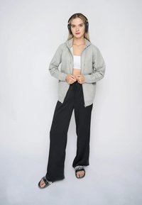 Siva zip-up hoodie s kapuljačom, nosi se preko bijelog kroptopa, u kombinaciji s crnim hlačama širokih nogavica i crnim sandalama s bijelim detaljima.