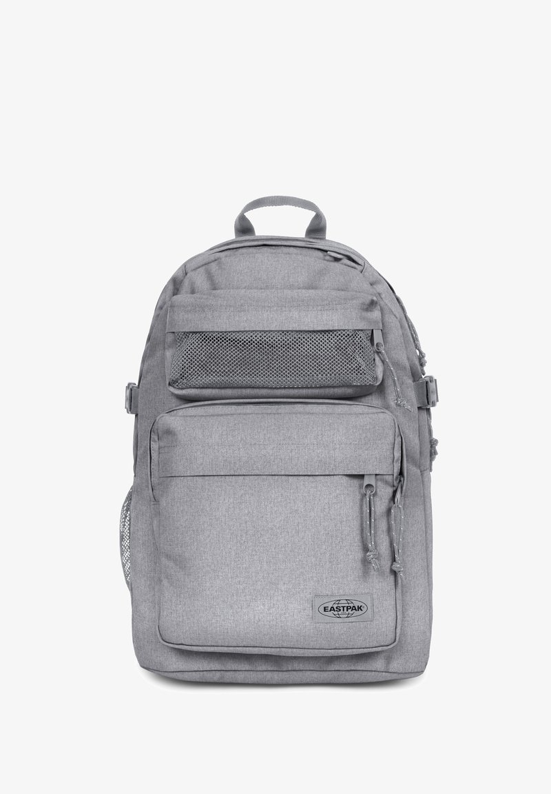 Mochila de tela gris con correas ajustables, bolsillos frontales y detalles de malla. Presenta cierres de cremallera y un parche con el logo.