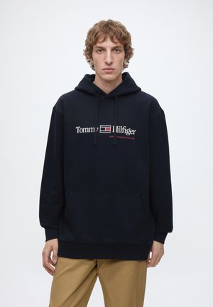Junger Mann mit lockigem Haar, der einen schwarzen Tommy Hilfiger Hoodie und beige Hosen trägt, steht vor einem schlichten weißen Hintergrund.