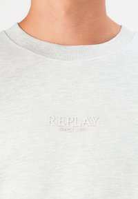 Light gray sweatshirt with a ribbed crew neck, featuring an embossed logo "REPLAY" and "SINCE 1981" on the chest. Soft texture.