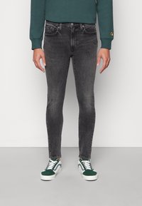 Zwarte skinny jeans met een lichte vervaging, vijfzakkenontwerp en een metalen knoopsluiting, gecombineerd met groene en witte sneakers.
