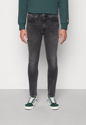 Jeans ajustados negros con un ligero desgaste, diseño de cinco bolsillos y cierre de botón metálico, combinados con zapatillas verdes y blancas.