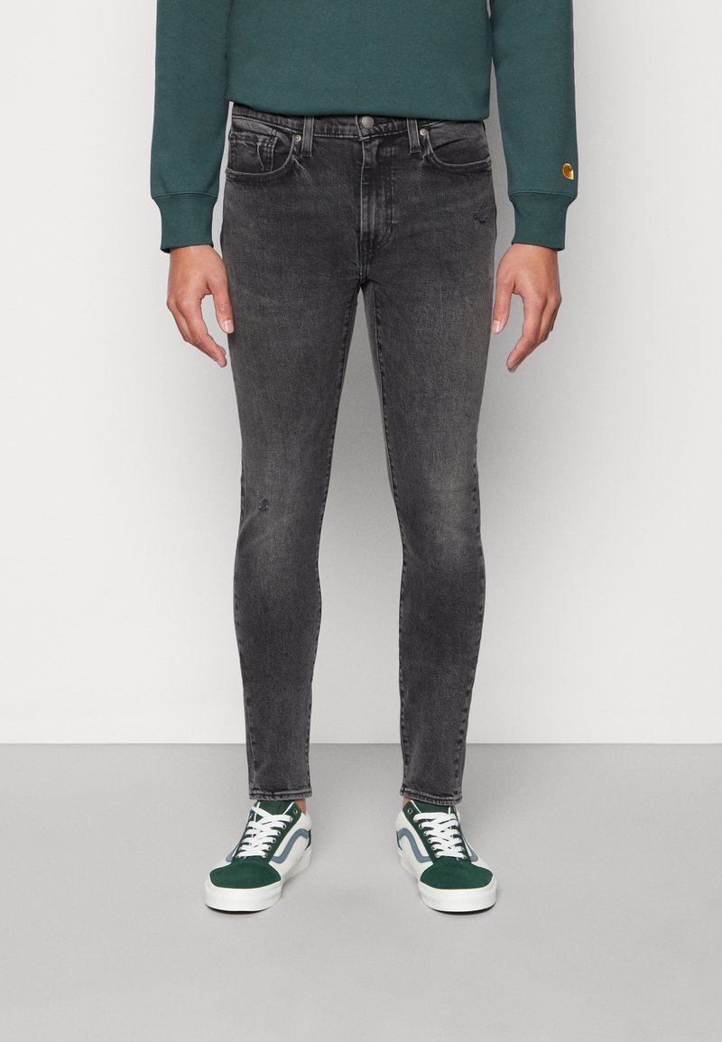 Zwarte skinny jeans met een lichte vervaging, vijfzakkenontwerp en een metalen knoopsluiting, gecombineerd met groene en witte sneakers.