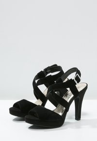 Svarta mockaklackar med korsade remmar, öppen tå, justerbar ankelspänne och strukturerade sulor för grepp.