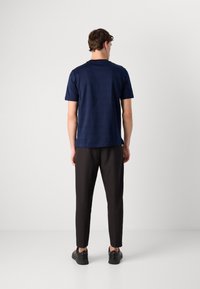 Homem de costas, vestindo uma camisa de manga curta azul marinho, calças pretas e ténis pretos, em frente a um fundo cinza claro liso.