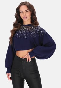 Pulover scurt de culoare bleumarin, cu aplicații de paiete sferice la guler, textură canalată și mâneci lungi, tip balon. Asortat cu pantaloni negri.