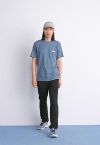 Mann som går innendørs iført en blå t-skjorte, svarte bukser, blå og hvite sneakers, og en grå caps foran en ensfarget hvit vegg.