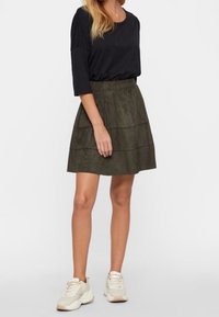 Sukně z olivově zeleného nubuku s vrstvenými díly, spárovaná s černým topem a světlými teniskami. Sukně má přiléhavý pas a rozšířený siluetu.