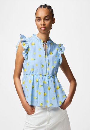 Femme aux cheveux tressés portant un haut bleu clair sans manches avec des manches à volants et un imprimé citron jaune, associé à un pantalon blanc.