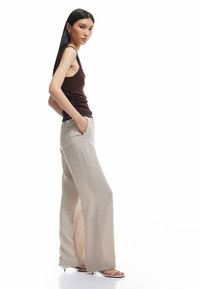 Brun, struktureret tanktop parret med lysebeige, brede bukser med lommer og et subtilt stribet mønster. Modellen står sidelæns.