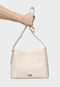 Bolso tote beige con un patrón texturizado, parte superior curva y correa ajustable, con un logo dorado de "TOUS" en la parte inferior.