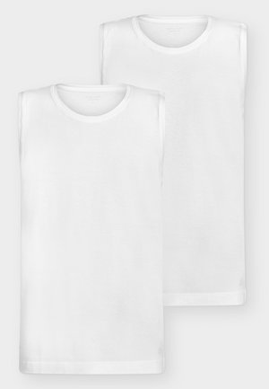 Uomo con capelli ricci rossi che indossa una giacca beige con bottoni sopra una camicia bianca, pantaloni crema e scarpe di pelle marroni, in piedi su uno sfondo bianco.