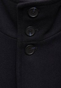 Detalle de un abrigo de lana oscuro que presenta un cuello alto, tres botones negros con cuatro orificios y una textura suave con costuras limpias.