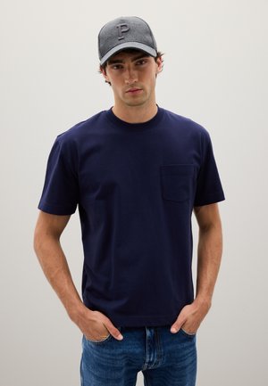 Camiseta de algodón azul marino con cuello redondo y mangas cortas, que presenta un bolsillo en el pecho. Llevada con jeans de denim azul claro y una gorra gris.